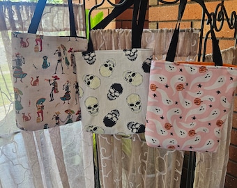 May include: Three tote bags with black handles hang on a black metal rack. The bags feature Halloween-themed patterns: a Day of the Dead design, skulls, and ghosts with pumpkins. The bags have black handles and are made of fabric.