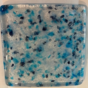 May include: Square glass art piece with a mosaic design. The artwork features a clear base with embedded blue and dark blue glass fragments. The glass has a slightly textured surface and a bubbly appearance.