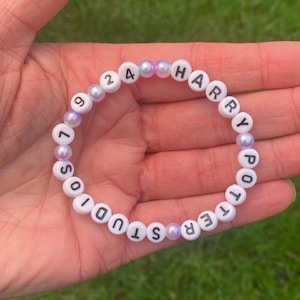 May include: White beaded bracelet with the words "Harry Potter Studios" and the numbers "1924" spelled out. The beads are interspersed with iridescent purple and white beads.