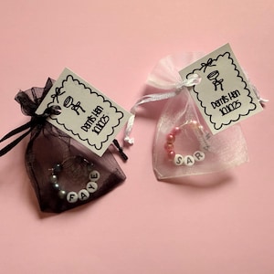 May include: Two small organza bags, one black and one white, each containing a beaded bracelet. The black bag has a bracelet spelling "FAYE", and the white bag spells "SAR". Each bag has a tag with a drawing and text.