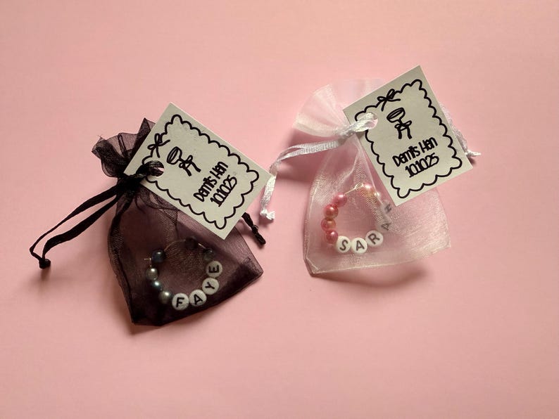 May include: Two small organza bags, one black and one white, each containing a beaded bracelet. The black bag has a bracelet spelling "FAYE", and the white bag spells "SAR". Each bag has a tag with a drawing and text.