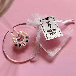 May include: A champagne glass with a beaded ring spelling "BRIDE" and a small gift bag with a tag that reads "Demi's Hen 10.10.25" on a pink background. The ring is made of white beads and the letters are black.