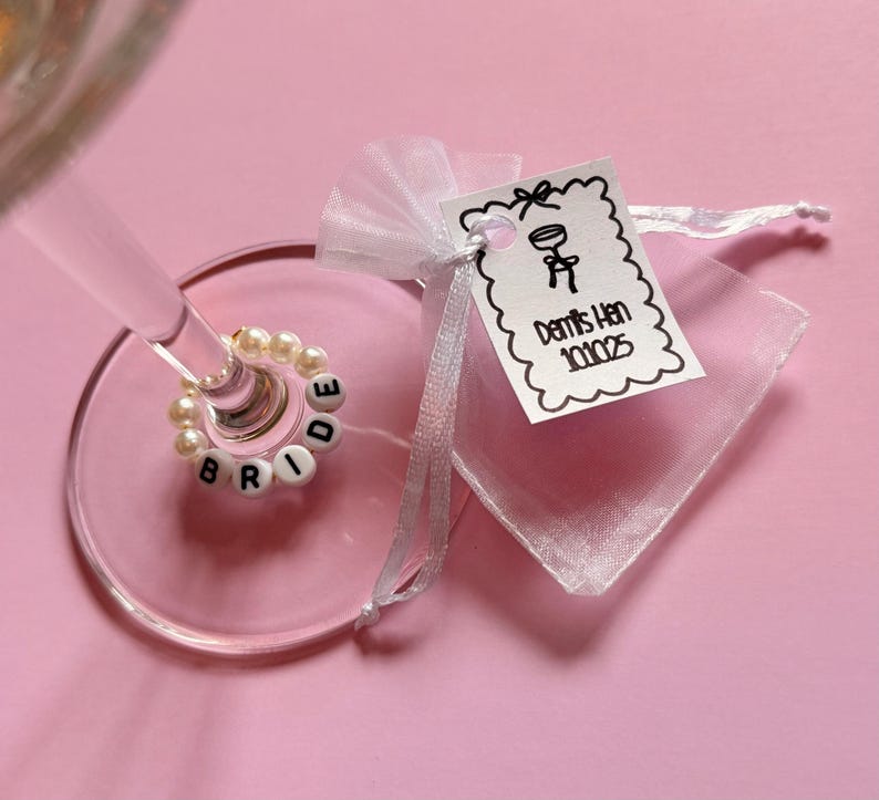 May include: A champagne glass with a beaded ring spelling "BRIDE" and a small gift bag with a tag that reads "Demi's Hen 10.10.25" on a pink background. The ring is made of white beads and the letters are black.