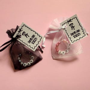 May include: Two small gift bags with personalized beaded bracelets inside. The bags are made of sheer fabric and have tags attached that say "Demis Han 101025". One bag is black and the other is white.
