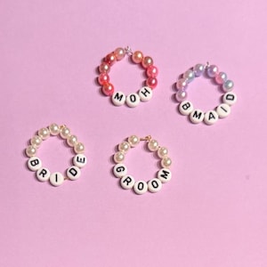 May include: Four beaded bracelets with white letter beads spelling out "BRIDE", "GROOM", "MOH", and "MAID". The bracelets feature pearls in various colors, including pink, peach, and white, set against a pink background.