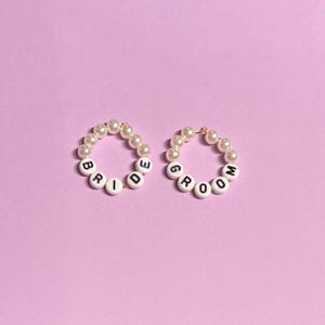 May include: Two beaded bracelets with the words "BRIDE" and "GROOM" spelled out in white letter beads. The bracelets are made of small, cream-colored pearls and are set against a light purple background.