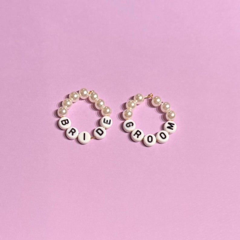 May include: Two beaded bracelets with the words "BRIDE" and "GROOM" spelled out in white letter beads. The bracelets are made of small, cream-colored pearls and are set against a light purple background.