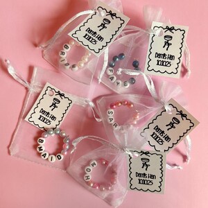 May include: Small white organza bags containing beaded bracelets with names like "Bride", "Maid", "Sarah", and "MOH". Each bag has a tag that says "Demi's Hen 101025" and a small graphic. The background is a solid pink.