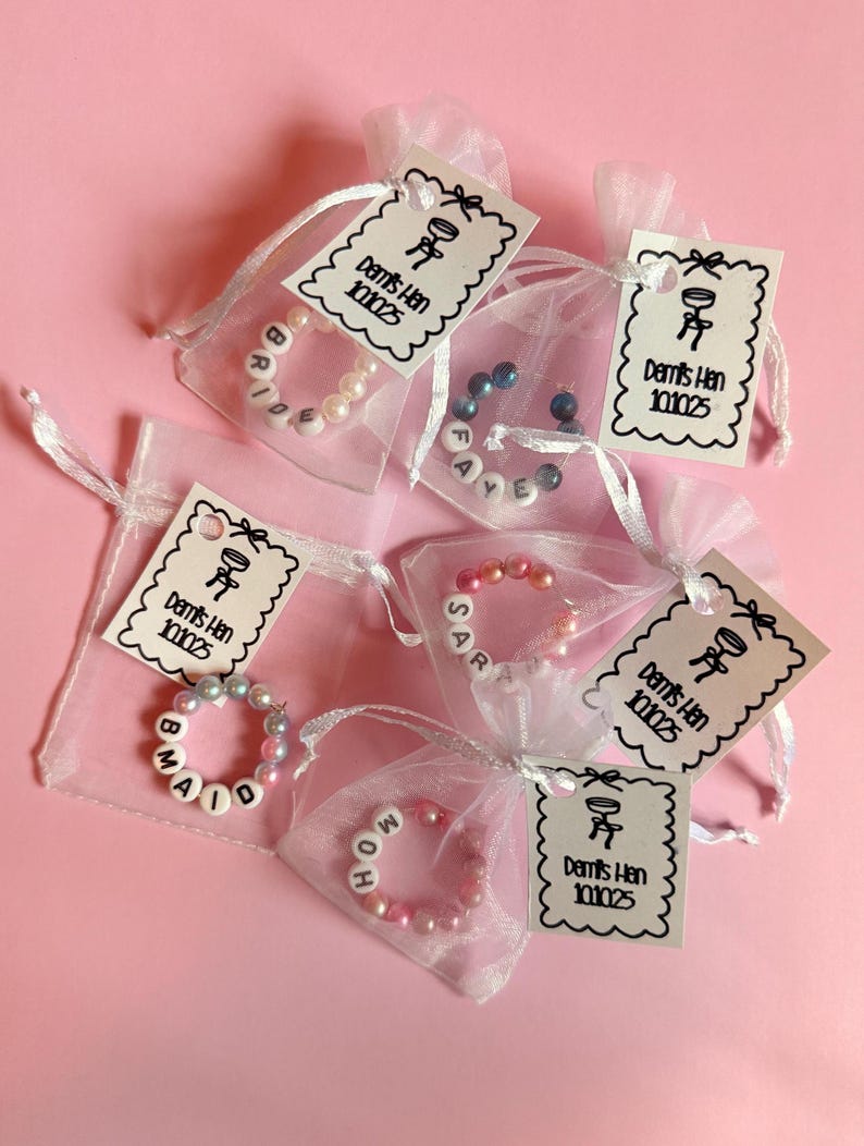 May include: Small white organza bags containing beaded bracelets with names like "Bride", "Maid", "Sarah", and "MOH". Each bag has a tag that says "Demi's Hen 101025" and a small graphic. The background is a solid pink.