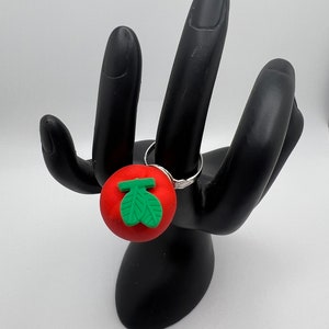 May include: A silver ring with a red, round charm featuring a green leaf design. The ring is displayed on a black hand-shaped jewelry holder against a white background. The charm is approximately 1 inch in diameter.