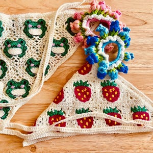 May include: Handmade crochet headbands. One headband features green frog faces, the other has red strawberries. A floral embellishment with pink and blue flowers adorns the strawberry headband. The headbands are cream-colored and made of yarn.