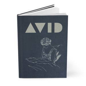 May include: A hardbound journal with a dark blue cover. The word "AVID" is printed in a light gray font at the top. Below the text is a line drawing of a person reading. The journal is open to reveal white pages.