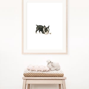 May include: A framed watercolor print of a Boston Terrier dog in black and white, playing with a small object. The print is in a light wood frame. Below the print, a stack of blankets and a stuffed animal are on a wooden stool.