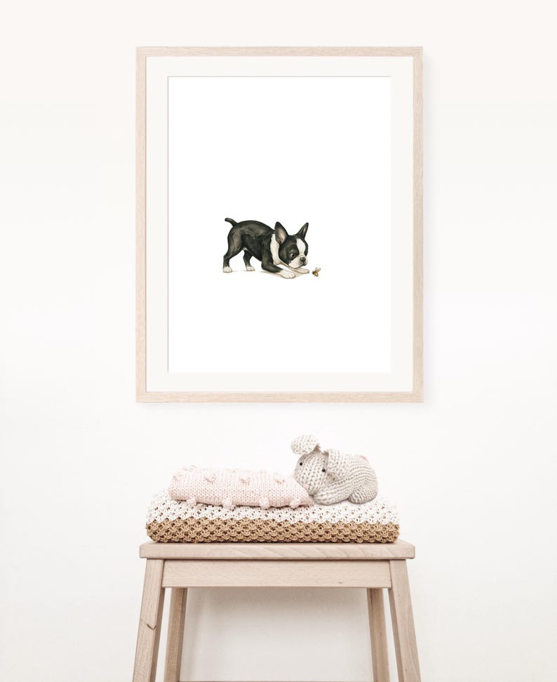 May include: A framed watercolor print of a Boston Terrier dog in black and white, playing with a small object. The print is in a light wood frame. Below the print, a stack of blankets and a stuffed animal are on a wooden stool.