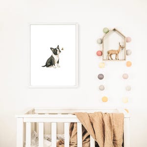May include: A framed watercolor print features a black and white Boston Terrier puppy gazing at a small bee. The artwork is set against a white background, with a white frame. The print is displayed above a white crib with a tan blanket.