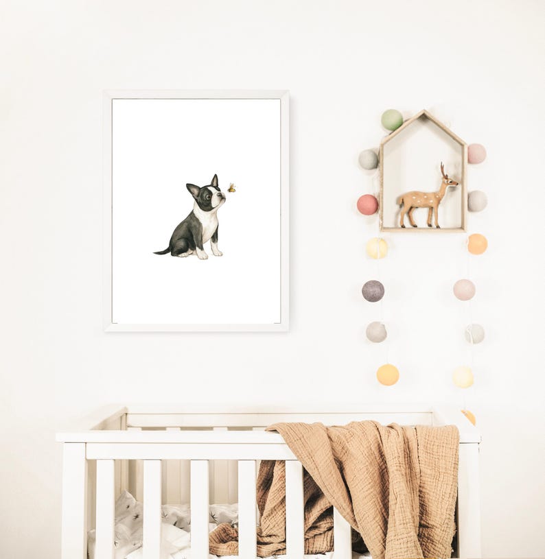 May include: A framed watercolor print features a black and white Boston Terrier puppy gazing at a small bee. The artwork is set against a white background, with a white frame. The print is displayed above a white crib with a tan blanket.