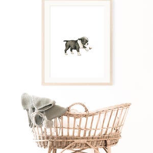 May include: A framed watercolor print of a Boston Terrier puppy playing with a small object. The puppy is black and white, with a light brown object in its paw. Below the print is a wicker bassinet with a gray blanket.