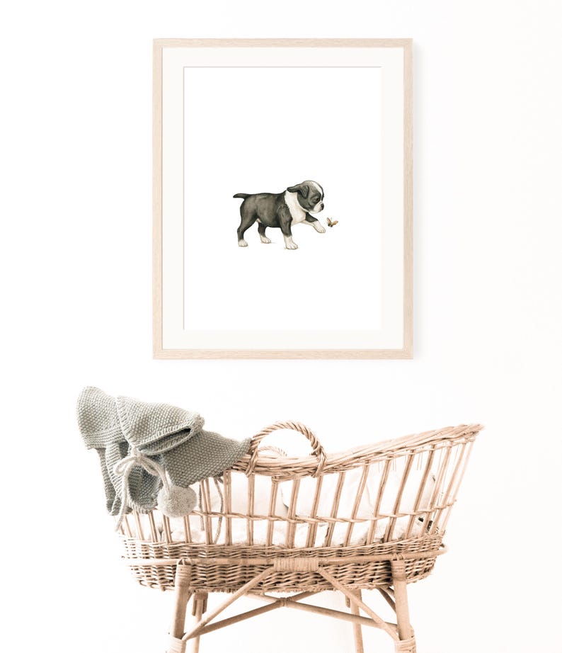 May include: A framed watercolor print of a Boston Terrier puppy playing with a small object. The puppy is black and white, with a light brown object in its paw. Below the print is a wicker bassinet with a gray blanket.