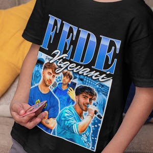May include: Black t-shirt featuring a graphic of a singer with the text "FEDE Vigevani" in blue. The design includes images of the singer in various poses. The shirt is worn by a child.