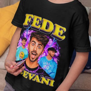 May include: Black t-shirt featuring a graphic of a man with the name "FEDE EVANI" in yellow lettering. The graphic includes images of the man and other people. The shirt is being worn by a child.