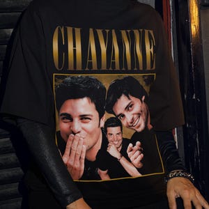 May include: Black t-shirt featuring the name "CHAYANNE" in gold lettering above a photo of three men. The t-shirt is worn by a person wearing black sunglasses and black clothing.