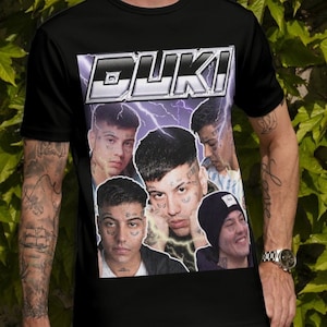 May include: Black t-shirt featuring a graphic of multiple faces and the word "DUKI" in a white and purple design. The shirt is worn by a person with tattoos and glasses.