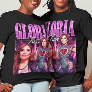 May include: Two people wearing black t-shirts with a pink and white graphic print of Gloria Trevi. The graphic features a photo of Gloria Trevi and the text "Gloria Trevi". The people are wearing blue jeans.