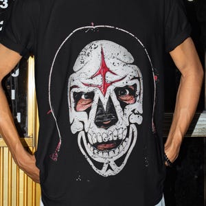 以下が含まれることがあります： 白と赤の頭蓋骨のデザインが特徴の黒いTシャツ。頭蓋骨にはフードと額に赤い星があります。サングラスとパールのネックレスを着用しています。背景は金と黒のドアです。