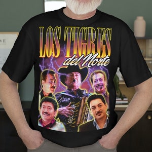 May include: A black t-shirt with a graphic print of a band called "Los Tigres del Norte". The graphic features five men, one playing an accordion, with lightning bolts and a purple and yellow color scheme.