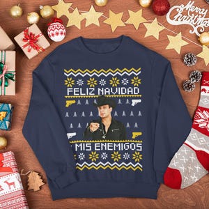 May include: Navy blue Christmas sweater with the text "Feliz Navidad Mis Enemigos" and a man pointing. The sweater has a festive pattern with yellow and white accents, and gun graphics. Surrounded by wrapped gifts and Christmas decorations.