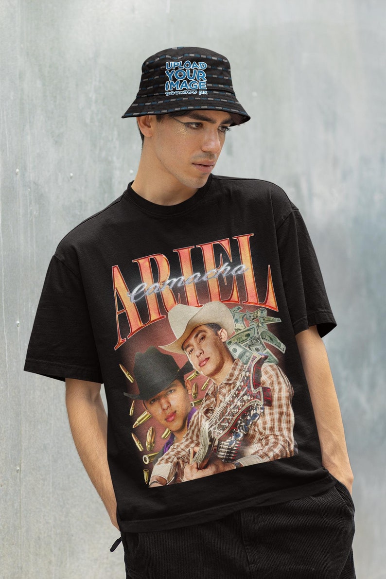Pu&ograve; includere: Una maglietta nera con una stampa grafica di due uomini con cappelli da cowboy. Gli uomini tengono in mano delle chitarre e sullo sfondo ci sono banconote e proiettili. La scritta "ARIEL Camacho" &egrave; stampata sulla maglietta.