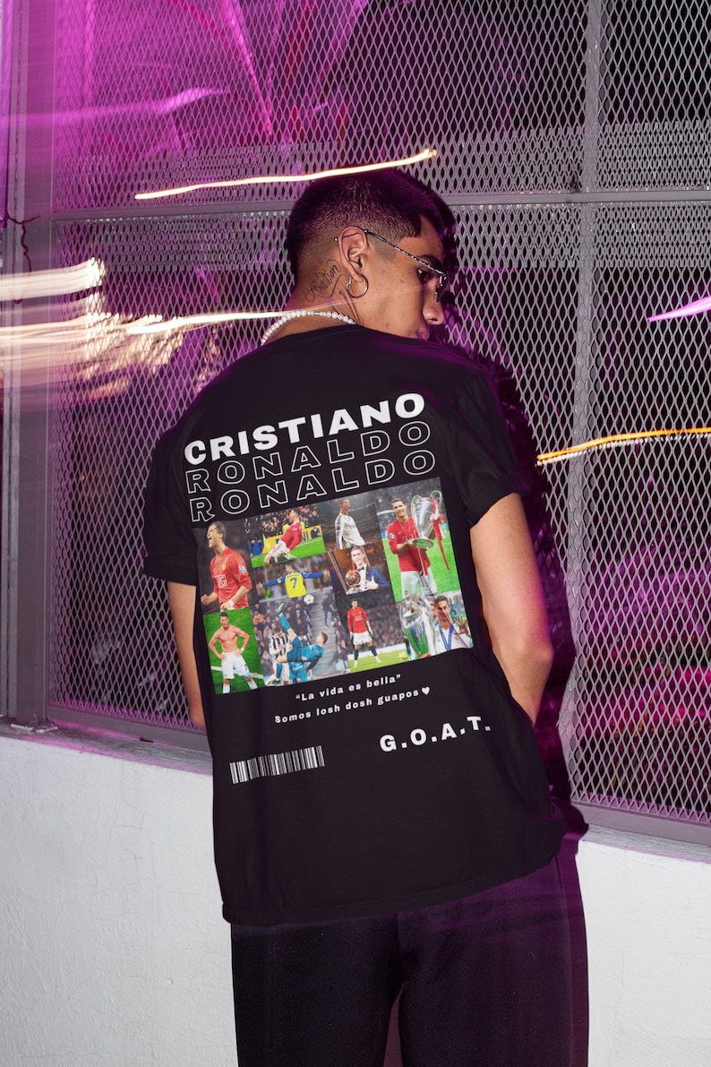 Pode incluir: Camiseta preta com uma colagem de imagens de Cristiano Ronaldo jogando futebol. O texto "CRISTIANO RONALDO" &eacute; impresso em branco nas costas da camiseta. O texto "La vida es bella" e "Somos losh dosh guapos" &eacute; impresso em branco abaixo da colagem. O texto "G.O.A.T." &eacute; impresso em branco abaixo do texto.