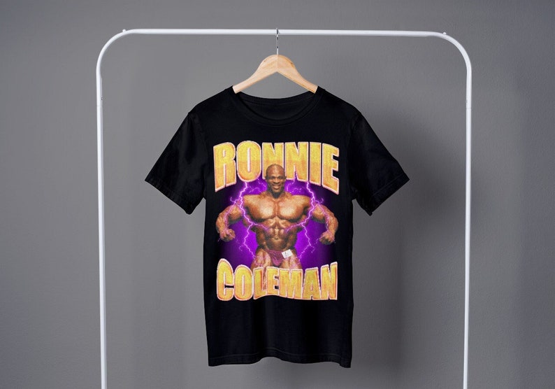 以下が含まれることがあります： 黄色い「RONNIE COLEMAN」の文字と稲妻が描かれた筋肉質の男性のグラフィックプリントが施された黒いTシャツです。