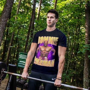 以下が含まれることがあります： 筋肉質の男性と「Donnie Coleman」という文字が描かれた黒いTシャツを着た人が、森の中でバーベルを持ち上げています。