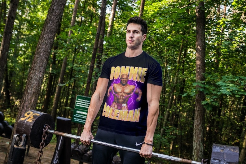 以下が含まれることがあります： 筋肉質の男性と「Donnie Coleman」という文字が描かれた黒いTシャツを着た人が、森の中でバーベルを持ち上げています。