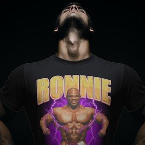 以下が含まれることがあります： 黄色のボディビルダーのグラフィックと黄色の文字で「RONNIE」と書かれた黒いTシャツ