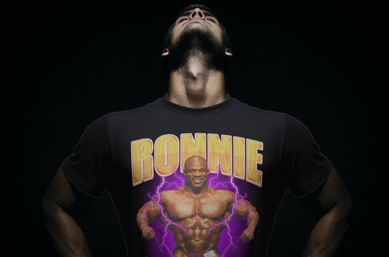 以下が含まれることがあります： 黄色のボディビルダーのグラフィックと黄色の文字で「RONNIE」と書かれた黒いTシャツ