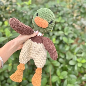 May include: A handmade crochet duck doll with a green head, brown wings, and an orange beak. The duck has a beige body and orange feet. The doll is held up against a background of green foliage.