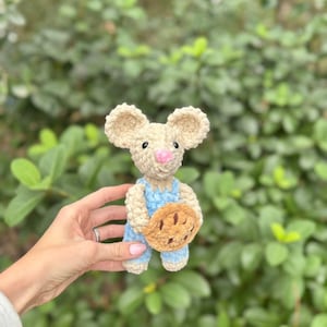 May include: A crocheted mouse figure with a light beige body, large ears, and a pink nose. It wears blue dungarees and holds a brown pie. The background is a blurred green bush.