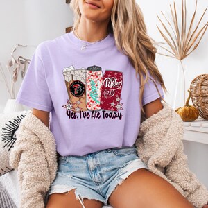 May include: A light purple t-shirt featuring a graphic of three beverages: a coffee drink, a soda can with a Christmas tree design, and a Dr. Pepper can. The text "Yes, I've Ate Today" is below the graphics. The shirt is worn by a person.