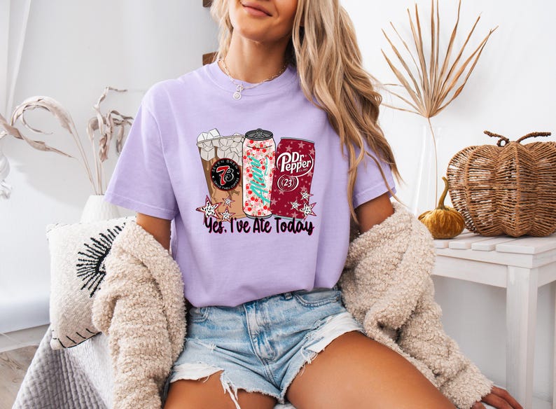 May include: A light purple t-shirt featuring a graphic of three beverages: a coffee drink, a soda can with a Christmas tree design, and a Dr. Pepper can. The text "Yes, I've Ate Today" is below the graphics. The shirt is worn by a person.