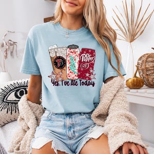 May include: Light blue t-shirt featuring three beverage designs: a coffee drink, a soda can with a Christmas tree, and a Dr. Pepper can. The text "Yes, I've Ate Today" is below the designs. The shirt is worn by a person.