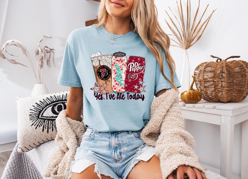 May include: Light blue t-shirt featuring three beverage designs: a coffee drink, a soda can with a Christmas tree, and a Dr. Pepper can. The text "Yes, I've Ate Today" is below the designs. The shirt is worn by a person.