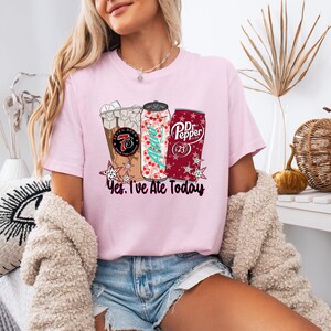 May include: Pink t-shirt with a graphic of a coffee drink, a soda can with a Christmas tree design, and a Dr. Pepper can. The text "Yes, I've Ate Today" is below the graphics. The shirt is worn by a person.