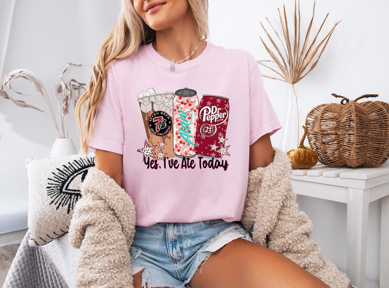 May include: Pink t-shirt with a graphic of a coffee drink, a soda can with a Christmas tree design, and a Dr. Pepper can. The text "Yes, I've Ate Today" is below the graphics. The shirt is worn by a person.
