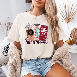 May include: Cream-colored t-shirt featuring a graphic of three beverages: an iced coffee, a soda can with a Christmas tree design, and a Dr. Pepper can. The text "Yes, I've Ate Today" is below the graphics.