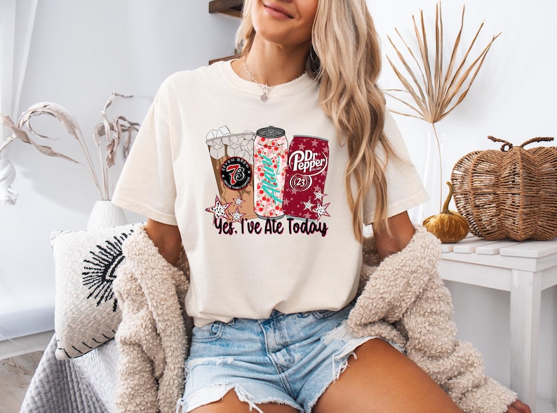 May include: Cream-colored t-shirt featuring a graphic of three beverages: an iced coffee, a soda can with a Christmas tree design, and a Dr. Pepper can. The text "Yes, I've Ate Today" is below the graphics.