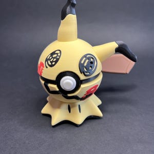 Op de afbeelding: Een 3D-geprint Mimikyu-figuur, een Pokémon-personage, in een staande pose. De figuur is voornamelijk geel met zwarte en rode accenten. Het heeft een lange, zwartgepunte aanhangsel en een kleine, bruinachtige vleugel. De basis is stervormig.