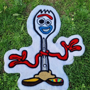 Può includere: Un tappeto di peluche bianco a forma del personaggio Forky dei film Toy Story. Forky è un forchettone di plastica bianco con occhietti tondi, un sorriso rosso e braccia rosse. Indossa scarpe blu e gialle.