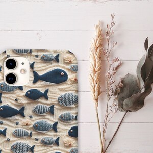 May include: A white phone case featuring a pattern of blue and white fish. The case has a camera lens cutout and a white background.
